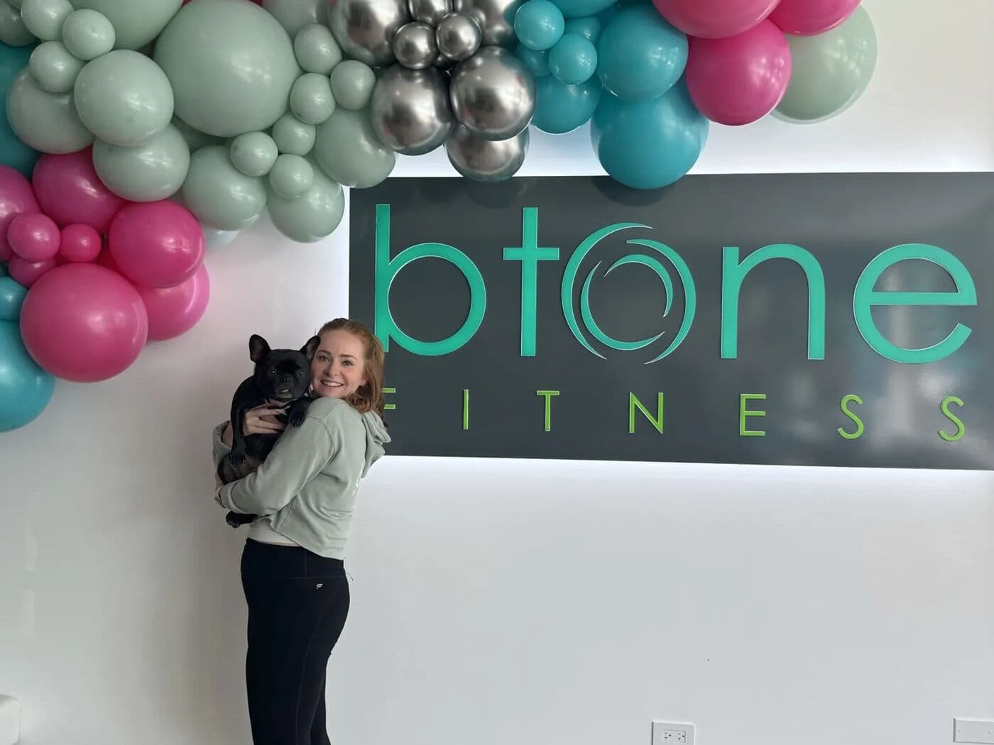 A woman holding a small dog in front of the btone FITNESS logo wall, decorated with balloons
