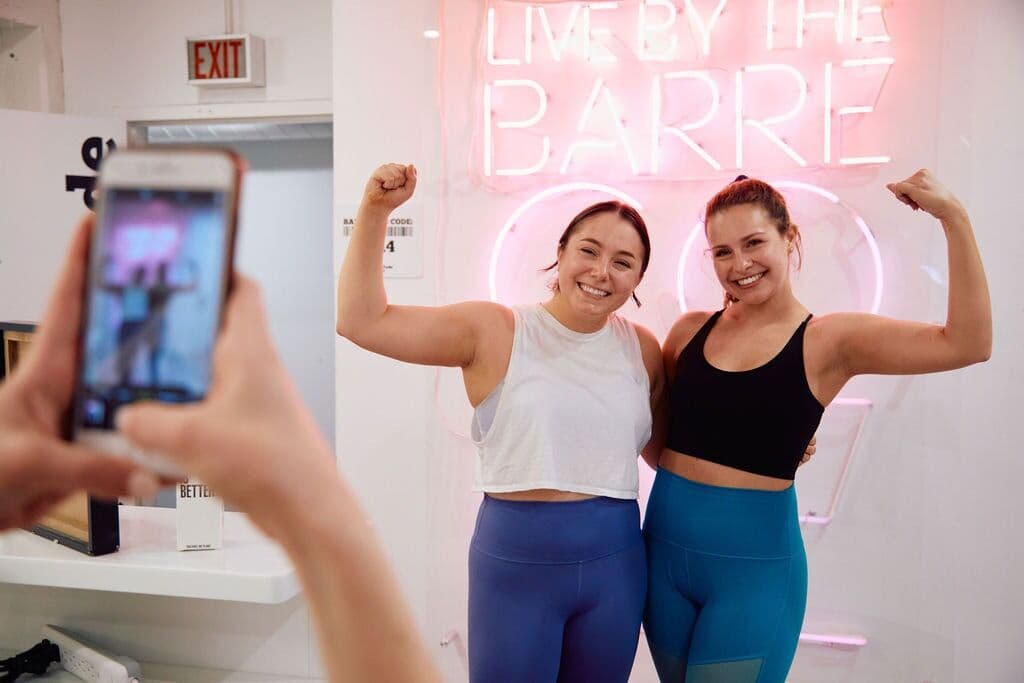 Two Barre Code instructors flexing in front of a neon sign while a friend takes their photo